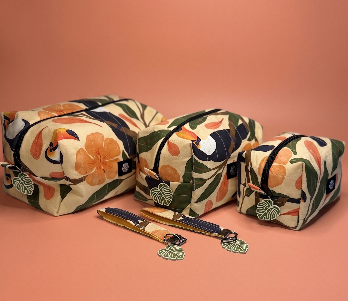 Three zippered cosmetic pouches with a tropical toucan and floral print in orange, green, and cream, displayed on a peach background with two matching keychains.