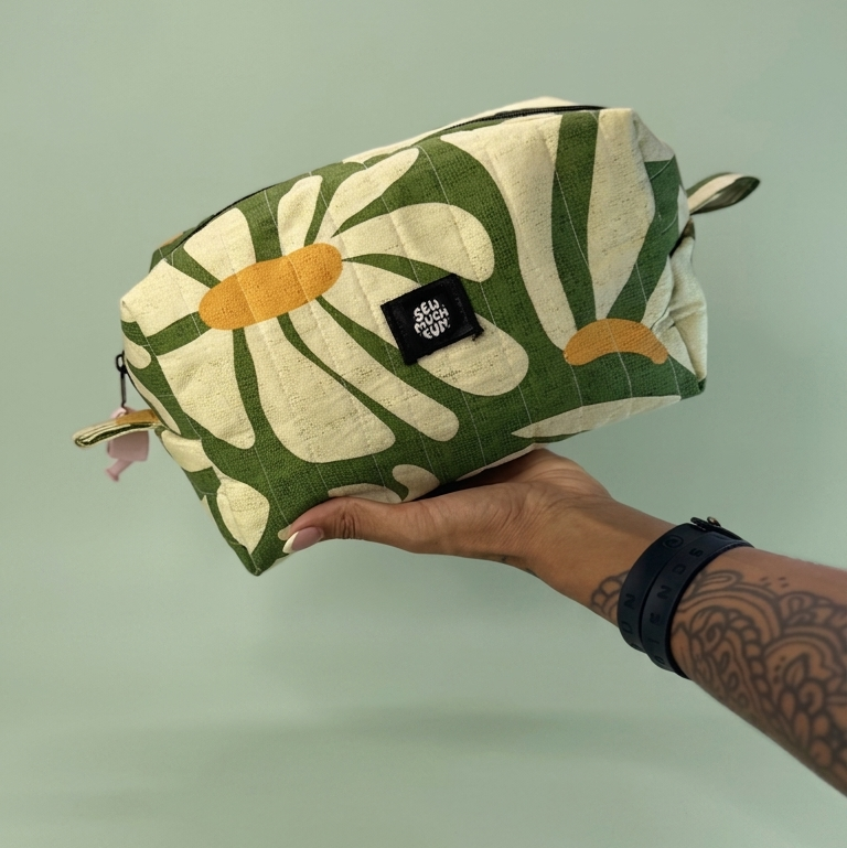 A hand holding a rectangular zippered pouch with a green and cream botanical pattern and orange accents, set against a solid pale green background.