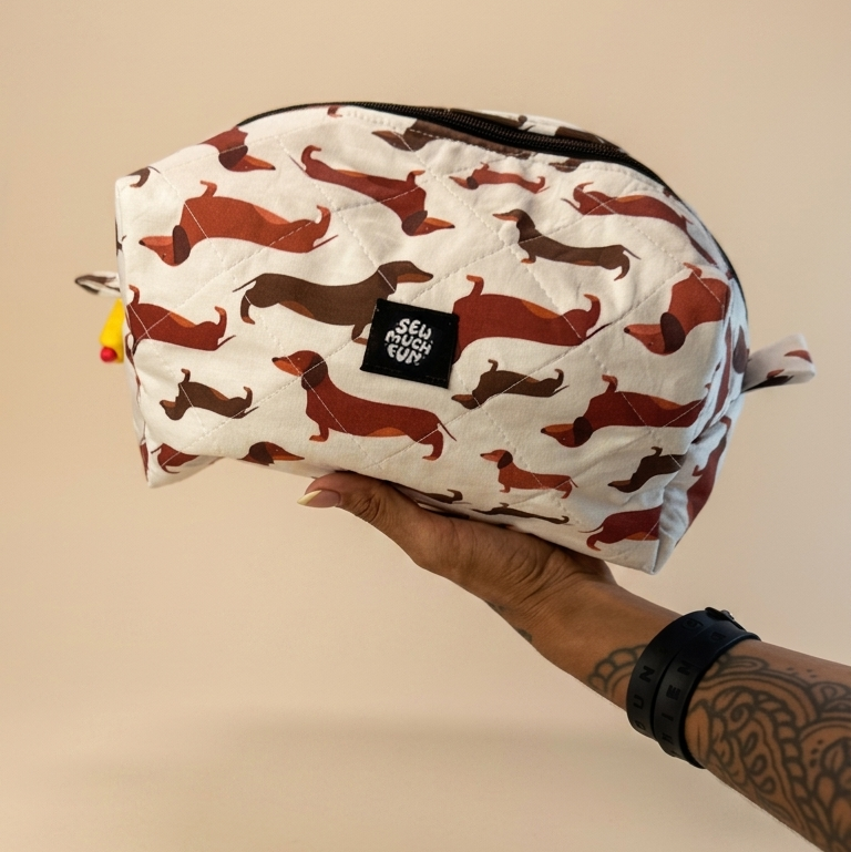 A hand holding a white zippered pouch decorated with a repeating pattern of brown dachshund dogs, set against a solid beige background.