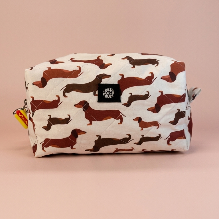 A rectangular zippered pouch with an allover dachshund print on an off-white background, featuring a small black square logo patch on the front and a small yellow tag on the left zipper pull, set against a solid dusty pink background.