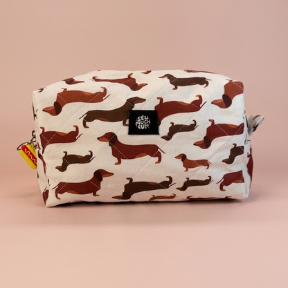 A rectangular zippered pouch with an allover dachshund print on an off-white background, featuring a small black square logo patch on the front and a small yellow tag on the left zipper pull, set against a solid dusty pink background.