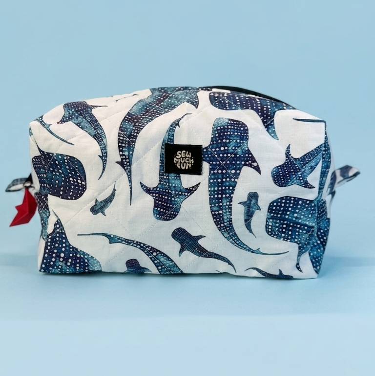 A rectangular zippered pouch with a white background and a blue indigo whale print, featuring a small black brand label on the front, set against a solid light blue background.