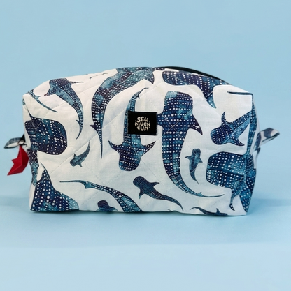 A rectangular zippered pouch with a white background and a blue indigo whale print, featuring a small black brand label on the front, set against a solid light blue background.