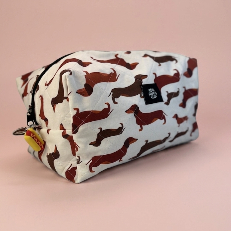 A rectangular zippered pouch with a white background and an all-over dachshund print in dark red and brown, shown on a solid light pink background.