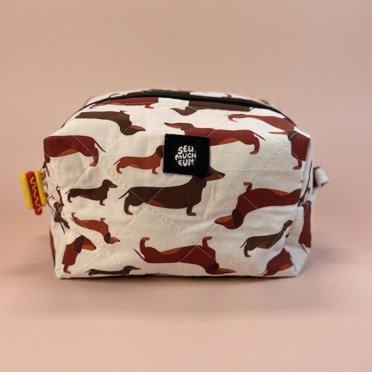A rectangular zippered pouch with an allover dachshund camouflage pattern in brown and black on a white background, featuring a black brand patch that reads 'SELF ASHLEY CRAFT'.