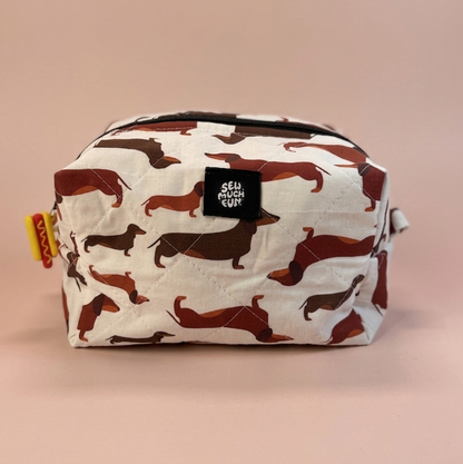 A rectangular zippered pouch with an allover dachshund camouflage pattern in brown and black on a white background, featuring a black brand patch that reads 'SELF ASHLEY CRAFT'.
