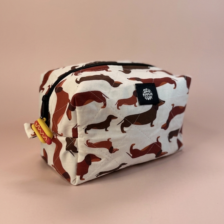 A rectangular zippered pouch with a white background and an allover print of brown dachshund dogs, featuring a small black brand tag on the front.