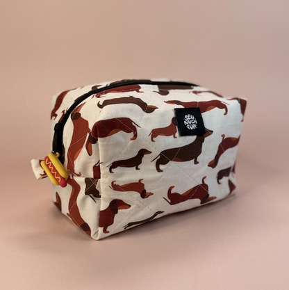 A rectangular zippered pouch with a white background and an allover print of brown dachshund dogs, featuring a small black brand tag on the front.
