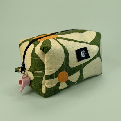 A rectangular fabric zip pouch with a retro floral pattern in olive green and cream, featuring a small pink teapot charm on the zipper pull and a black brand label.