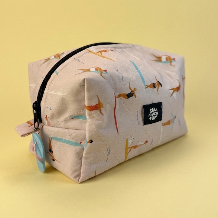 A rectangular zippered pouch with a light beige background and an all-over print of dachshunds in brown and orange, accented with light blue stripes. It features a black zipper, a small black brand label reading 'Dachshund Pups', and a small hanging tag on the left side, set against a solid pale yellow background.