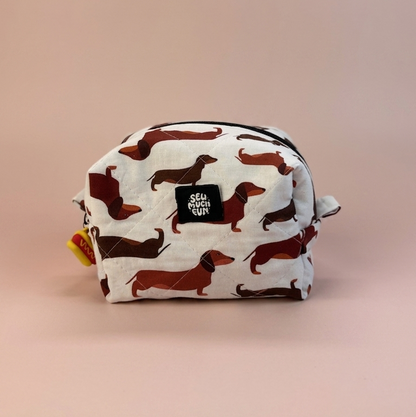 A small zippered pouch with an allover dachshund print on a white background, featuring a black 'GELLY PRINCE' brand patch on the front, set against a solid pale pink background.