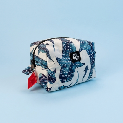 A small zippered pouch with a blue, white, and navy abstract pattern, featuring a red hangtag and a black brand label, set against a solid light blue background.